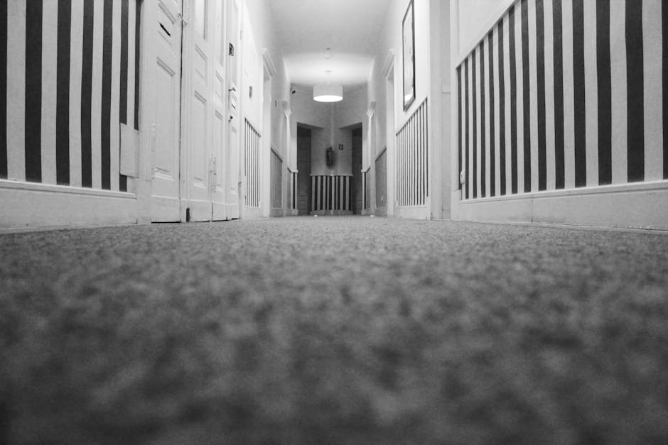 A low-angle, black-and-white image of a clean, textured carpeted hallway in a residential property. The perspective is from the floor, showing the carpet's surface and the striped wallpaper on both si